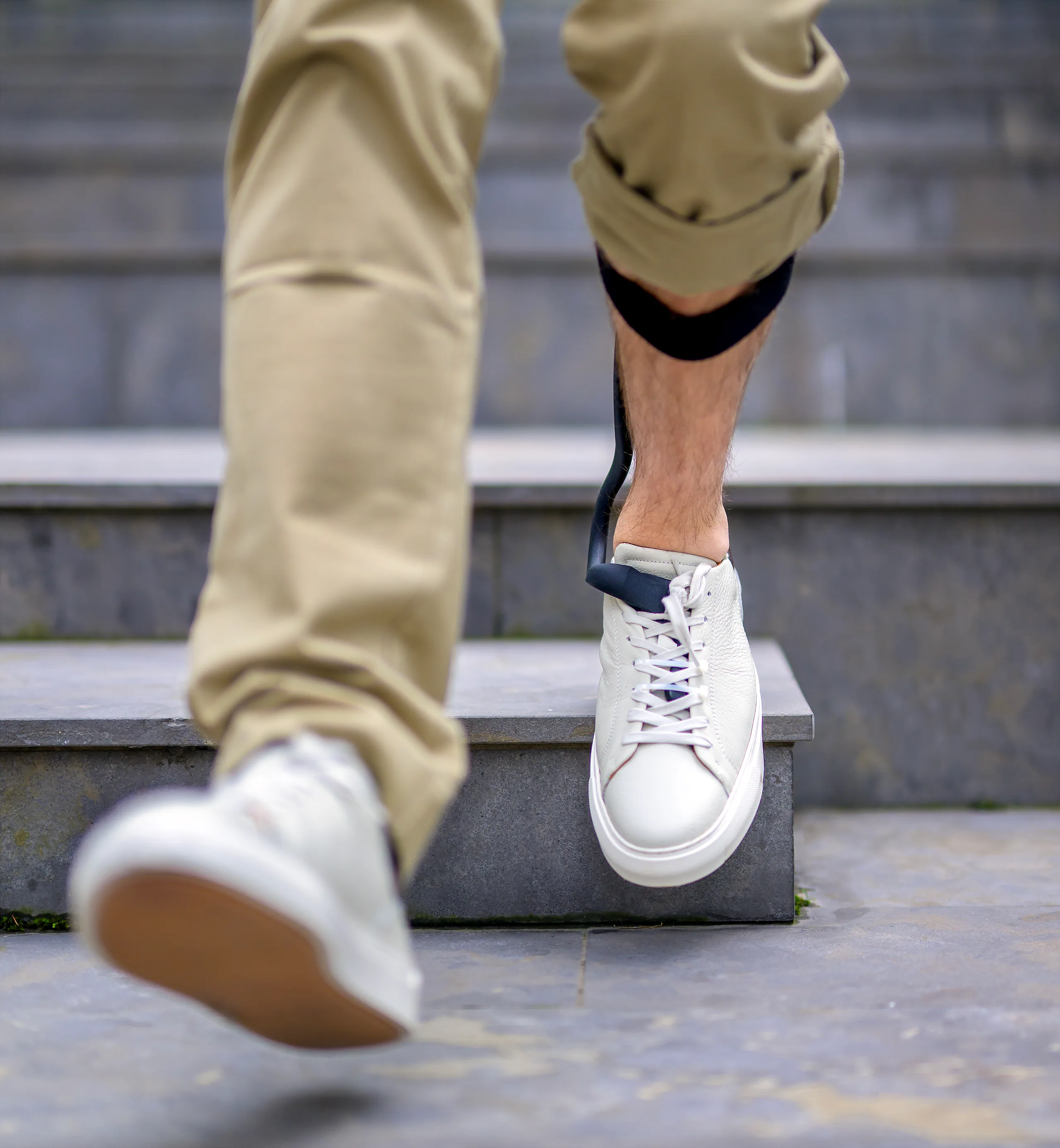 From behind: a man walks up a step. On his right lower leg is the Rise, with the strap below the knee and the thin strip down to the footplate under the laces of his white sneaker.
