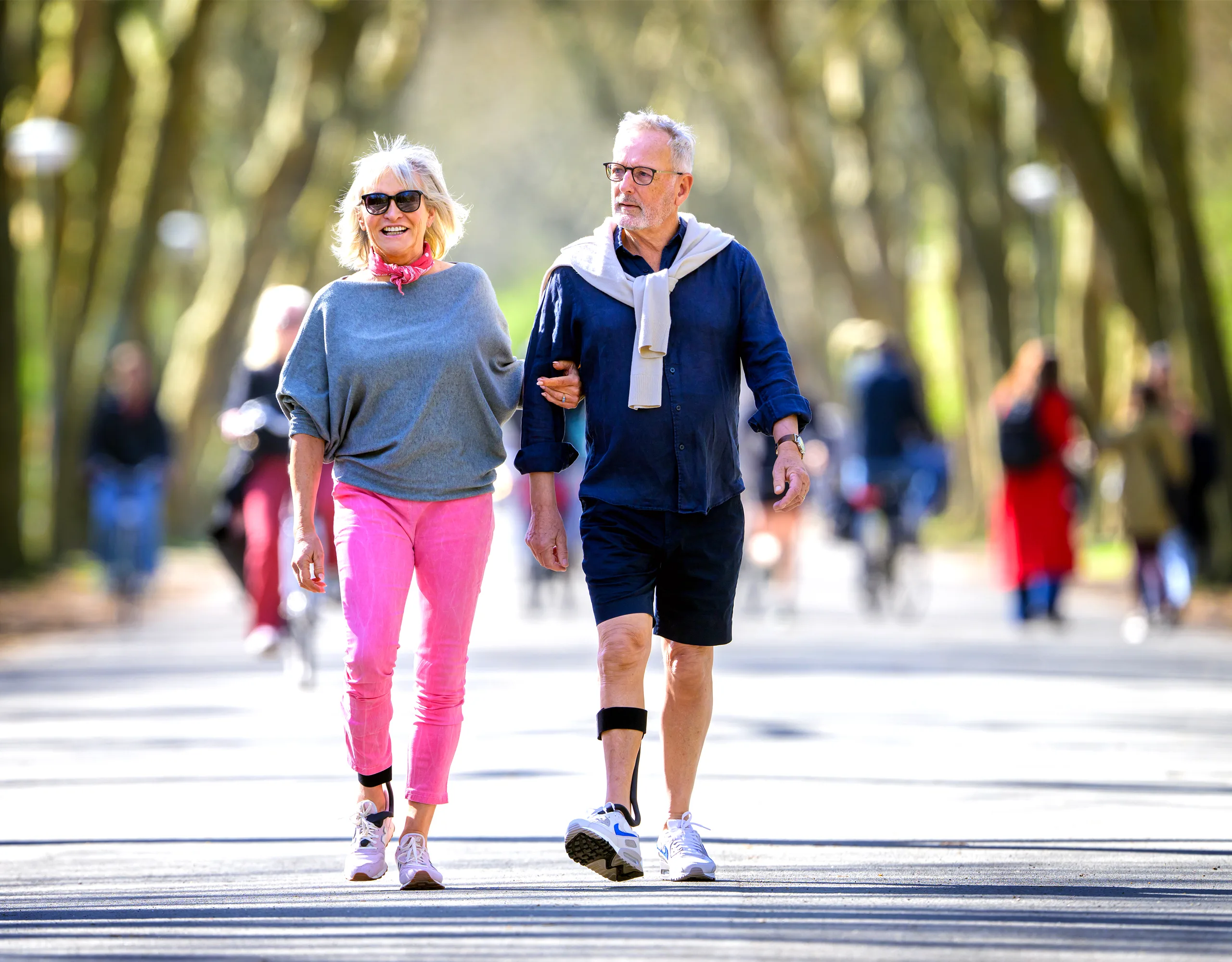 An older couple walking cheerfully arm-in-arm through a tree-lined park, the man wearing The Rise