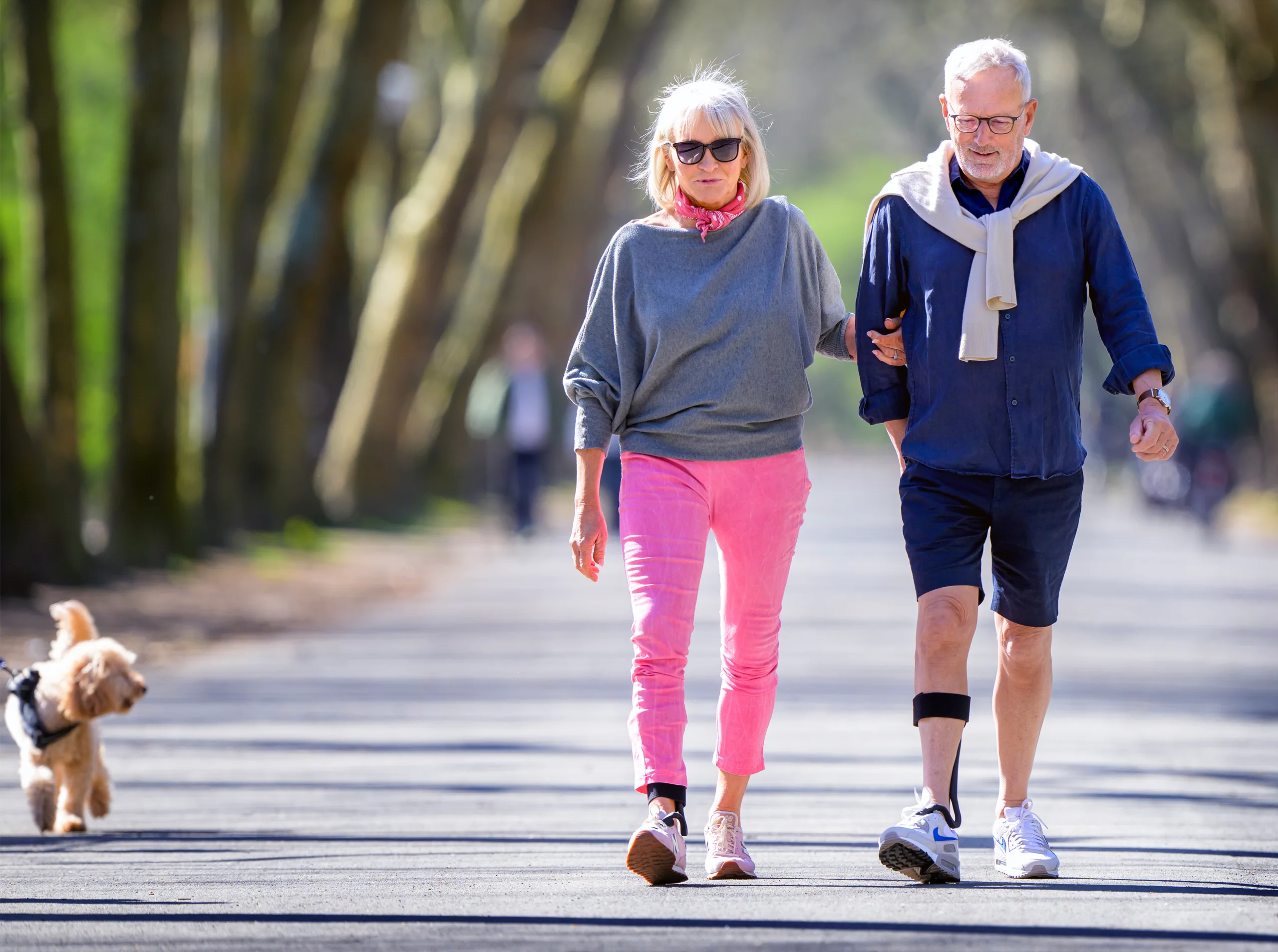 A woman and a man walk arm in arm along a park path. On his right lower leg is the Rise, with the strap below the knee.